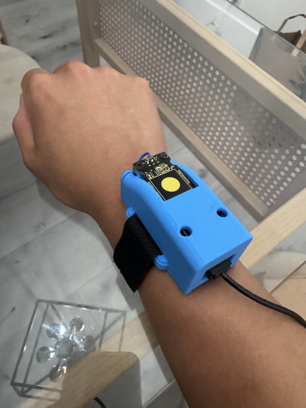Blue 3D-printed wristband housing strapped to a forearm with a velcro band, a capacitive touch module on top, and a tether cable leaving the end